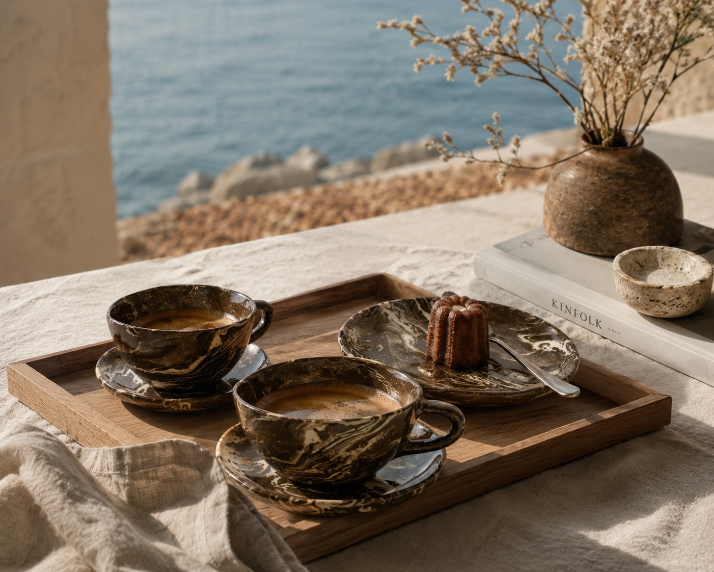 Ceramic cups and saucers on a wooden tray with a dessert, set against a scenic ocean view.