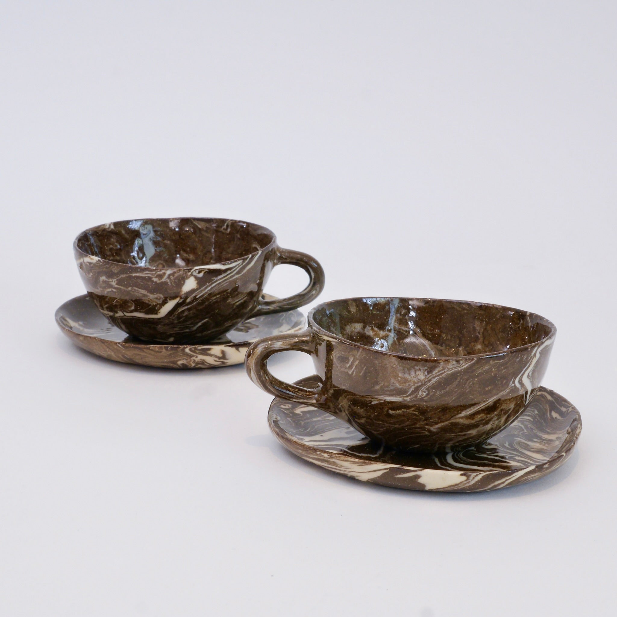 Two marbled brown ceramic cups with saucers on a light gray background